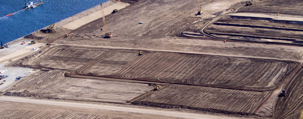 Aerial view of a construction site near a body of water, hinting at the expansion of Port Botany. The site features large, rectangular plots of dirt with visible tire tracks. Several cranes and construction vehicles are scattered throughout the area, working on land preparation for a future container terminal.