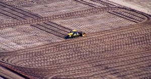 Aerial view of a large agricultural field with intricate tire track patterns; a lone tractor projects its vibrant color amidst earthy tones, diligently working at the center to cultivate the soil.