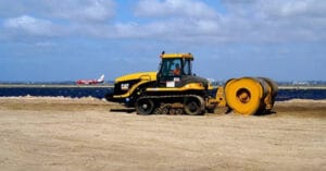 A yellow and black construction vehicle, ideal for various applications, is parked on a sandy area with a large cylindrical roller attached. In the background, a red and white airplane is visible against a cloudy sky.