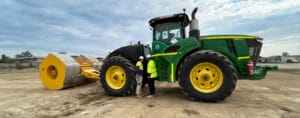 A large green tractor with yellow wheels is parked on a construction site under a cloudy sky. Two workers wearing hard hats and safety vests stand next to it, alongside a yellow cylindrical roller attachment, as they progress the Port Tanger development.