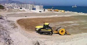 A construction site near the sea buzzes with activity as a tractor and roller work on a large circular dirt area, contributing to ambitious projects. In the background, cranes and unfinished buildings rise under a clear blue sky.