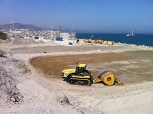 At Port Tanger, a yellow bulldozer operates at the construction site by the sea, leveling a circular patch of dirt. In the background of this project, there are cranes, stacked shipping containers, and a ship on the blue horizon. The sky is clear and sunny.