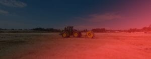 A tractor with a large yellow attachment drives across a barren dirt field under a deep blue sky, performing proof rolling. A red lens flare spreads from the right side, adding a dramatic effect to the scene. Trees and distant structures line the horizon.
