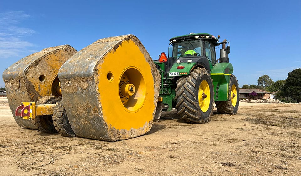 A green and yellow tractor on a dirt field is engaged in proof rolling, pulling two large hexagonal, yellow-colored rollers. The sky is clear and blue, with trees and a small building visible in the background.