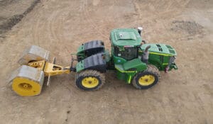 A green tractor is engaged in proof rolling on a dirt construction site, pulling a heavy roller. The machinery is parked on a vast, cleared earth surface, indicating land preparation or soil compaction work. The photo, captured from a high angle, highlights the intricacies of this essential process.
