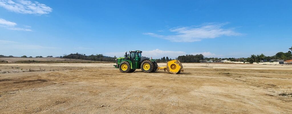 A green tractor with a yellow attachment is parked on a large, dusty field under a clear blue sky, ready for proof rolling. Trees and a few buildings are visible in the distant background.