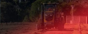 A large yellow tractor is smoothing a red dirt road with purpose and precision, surrounded by trees and a chain-link fence. The scene is bathed in a warm reddish glow, possibly due to lighting or effects, as if reviving land with each meticulous pass.