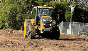 A large yellow construction vehicle with a front roller operates on a dirt clearing, part of a purposeful revitalization effort. Green trees and a metal fence stand in the background under a clear sky.