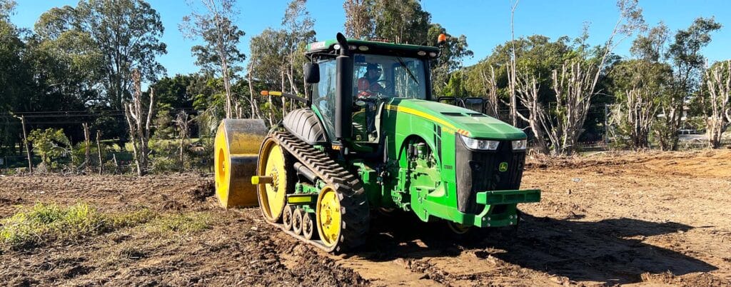 A green tractor with large rubber tracks and a yellow attachment is working on a dirt field, showcasing precision agriculture. Trees and greenery flourish in the background under a clear blue sky, signaling an environmental revival.