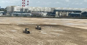 Two construction vehicles meticulously navigate a large, muddy field with visible tire tracks, showcasing applications in heavy-duty operations. In the background, industrial buildings and storage tanks stand tall, while red and white chimneys rise under a cloudy sky.
