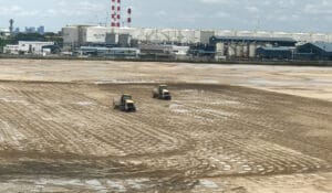 Two yellow excavators diligently work at the expansive, muddy Tuas Terminal construction site, marked by visible tire tracks. In the background, Phase 1 industrial buildings and storage tanks stand tall, accompanied by red and white chimneys under a cloudy sky.