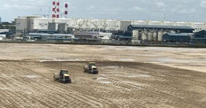 A barren field with two tractors diligently working the land under a cloudy sky, while in the background, an industrial complex projects its presence with large white buildings and multiple red-striped chimneys.