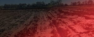The plowed field bears deep tire tracks, evidence of high energy impact compaction. Soil is turned for farming beneath a clear sky. Trees line the background, while a reddish hue gently blankets the right side of this serene image.