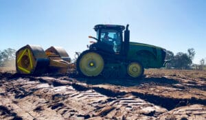 A green tractor rolls over a plowed field under a clear blue sky. With its large roller attachment, it applies high energy impact compaction to flatten the soil effectively. Deep tire tracks mark the ground, while trees stand visible in the background.