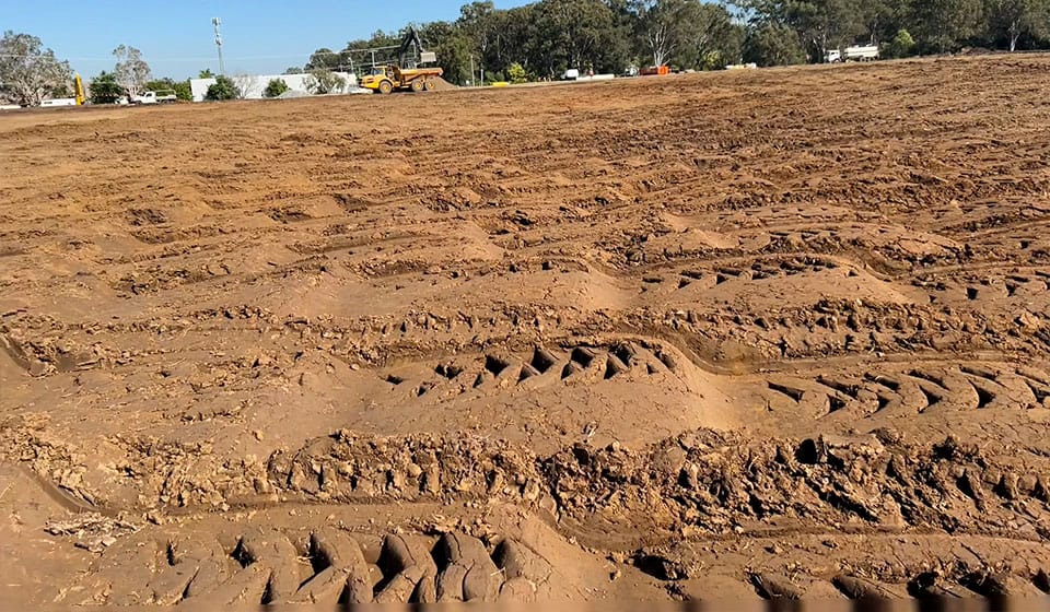 A muddy field reveals deep, crisscrossing tire tracks under a clear sky. In the distance, construction equipment is busy with high-energy impact compaction of placed fill, bordered by trees and buildings.