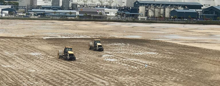 Two tractors work diligently on leveling a large, muddy field at Tuas Terminal, Phase 1, with the looming industrial complex in the background. The buildings have chimneys and industrial structures, evoking a factory setting. An overcast sky lends a muted tone to the scene.