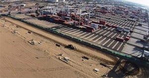 Aerial view of a bustling shipping port with numerous cranes and stacked shipping containers. In the foreground, construction projects are underway, with sandy areas dotted by equipment and vehicles. The horizon reveals a cityscape in the distance.