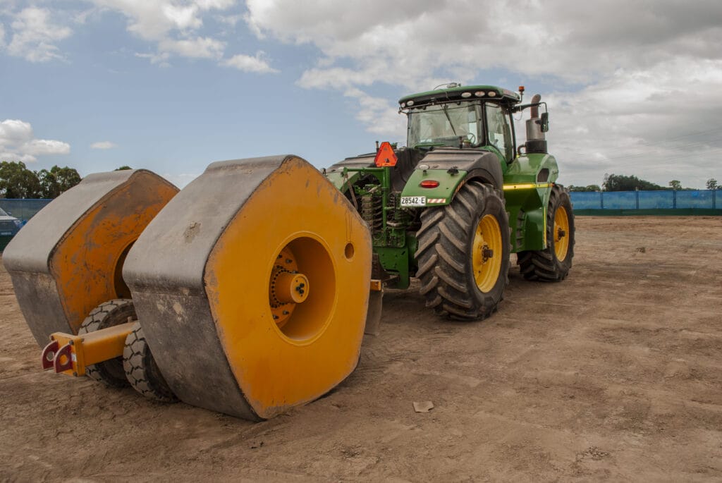 A green tractor with large rear tires is parked on a dirt surface. It is attached to a heavy, yellow dual-drum roller. The background features a blue fence and a partly cloudy sky.