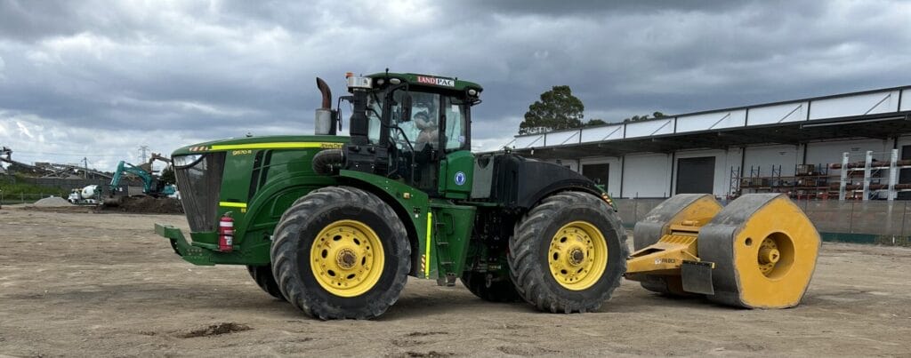 A green tractor with large yellow wheels is towing a heavy yellow roller on a construction site. The sky is overcast, and there are industrial buildings in the background. The ground appears to be dirt or gravel.