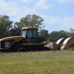 A yellow and black road roller, emblematic of sustainable construction practices, is parked on a grassy field. The machine's large cylindrical drum at the front efficiently compacts soil. Trees and a cloudy blue sky provide a serene backdrop to this scene.