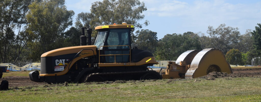 A yellow and black road roller, emblematic of sustainable construction practices, is parked on a grassy field. The machine's large cylindrical drum at the front efficiently compacts soil. Trees and a cloudy blue sky provide a serene backdrop to this scene.