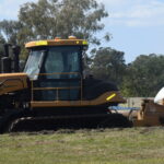 A yellow and black road roller, emblematic of sustainable construction practices, is parked on a grassy field. The machine's large cylindrical drum at the front efficiently compacts soil. Trees and a cloudy blue sky provide a serene backdrop to this scene.