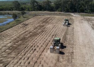 Two tractors work on a large, grid-patterned dirt field, creating parallel lines. The area is surrounded by trees and a small body of water, indicating a rural landscape under a clear sky.