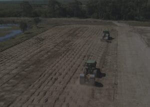 Two tractors are seen plowing a large, open field with rows of turned soil. The scene is set against a backdrop of trees and a small body of water on a clear day.