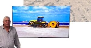 A person stands in front of a large TV screen displaying an image of a yellow construction vehicle on a sandy beach. The sky is blue with scattered clouds.