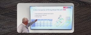 A man pointing at a presentation slide on a screen displaying information about in-situ compaction of mining mixed fills with HEIC, including a table and diagram. Background shows a textured surface pattern.