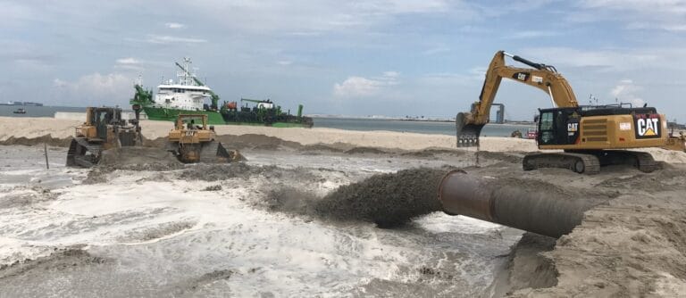 A construction site on a beach buzzes with heavy machinery, including an excavator and bulldozer, utilizing High Energy Impact Compaction for land reclamation. A large pipe discharges sand and water as a ship sails on the horizon under a cloudy sky.