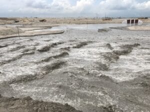 A construction site near water, featuring muddy ripples in the foreground. Heavy machinery for High Energy Impact Compaction and materials are visible in the background under a cloudy sky.