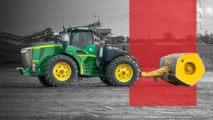 A green and yellow tractor with large tires is hitched to a heavy yellow compactor roller, set on a construction site. The background is grayscale, while the tractor and a red rectangle overlay are in color.