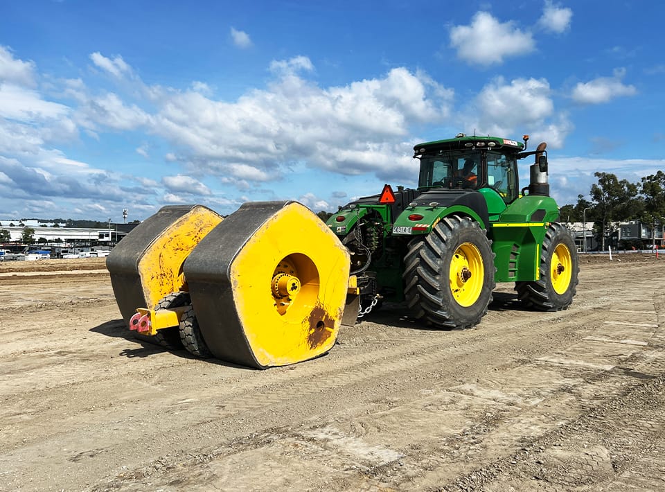 A green and yellow tractor pulls large yellow roller attachments over a dirt construction site under a partly cloudy sky. Buildings and trees are visible in the background.