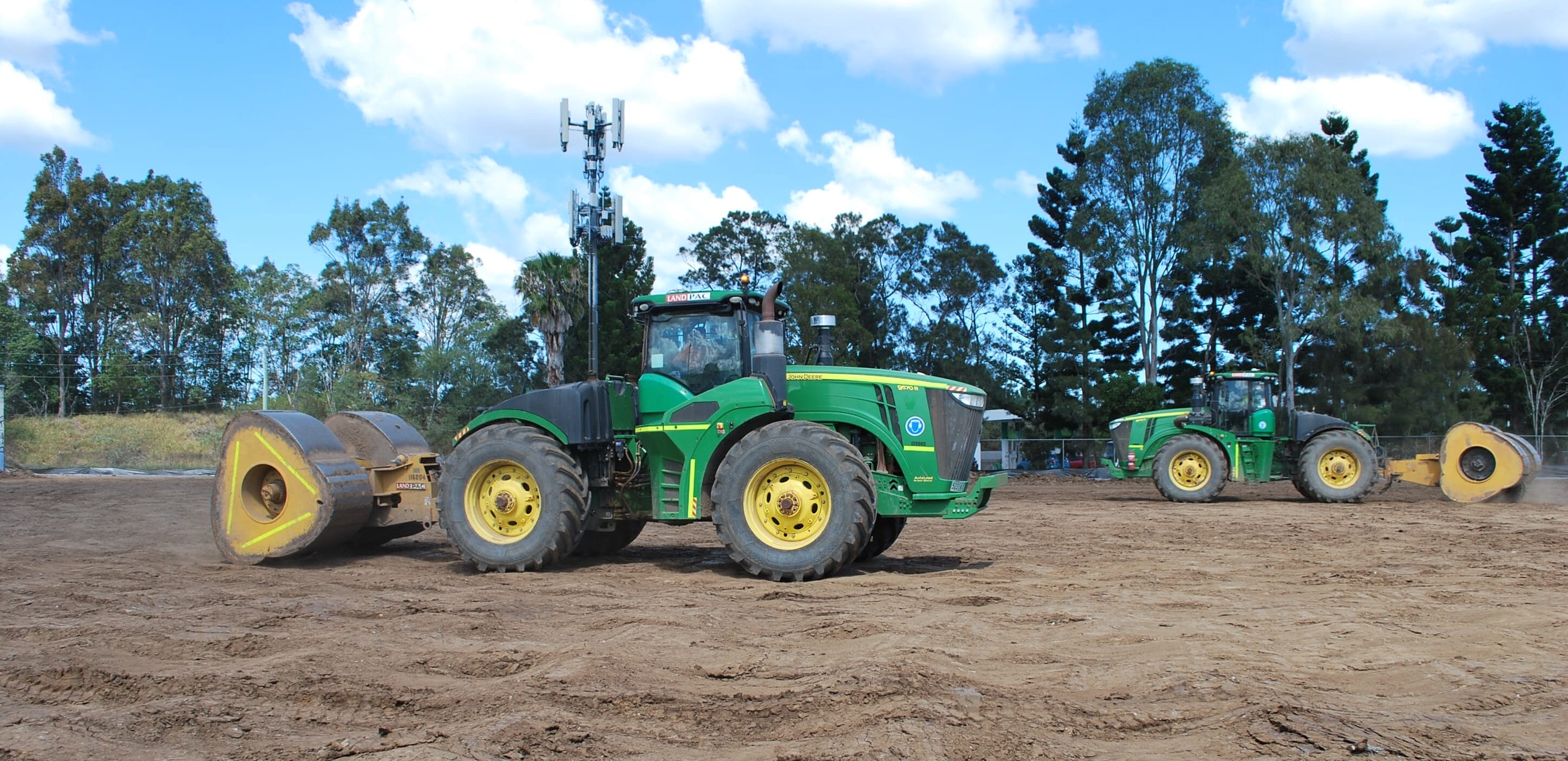 Two green tractors level contaminated fill on a dirt field, surrounded by trees and blue sky.