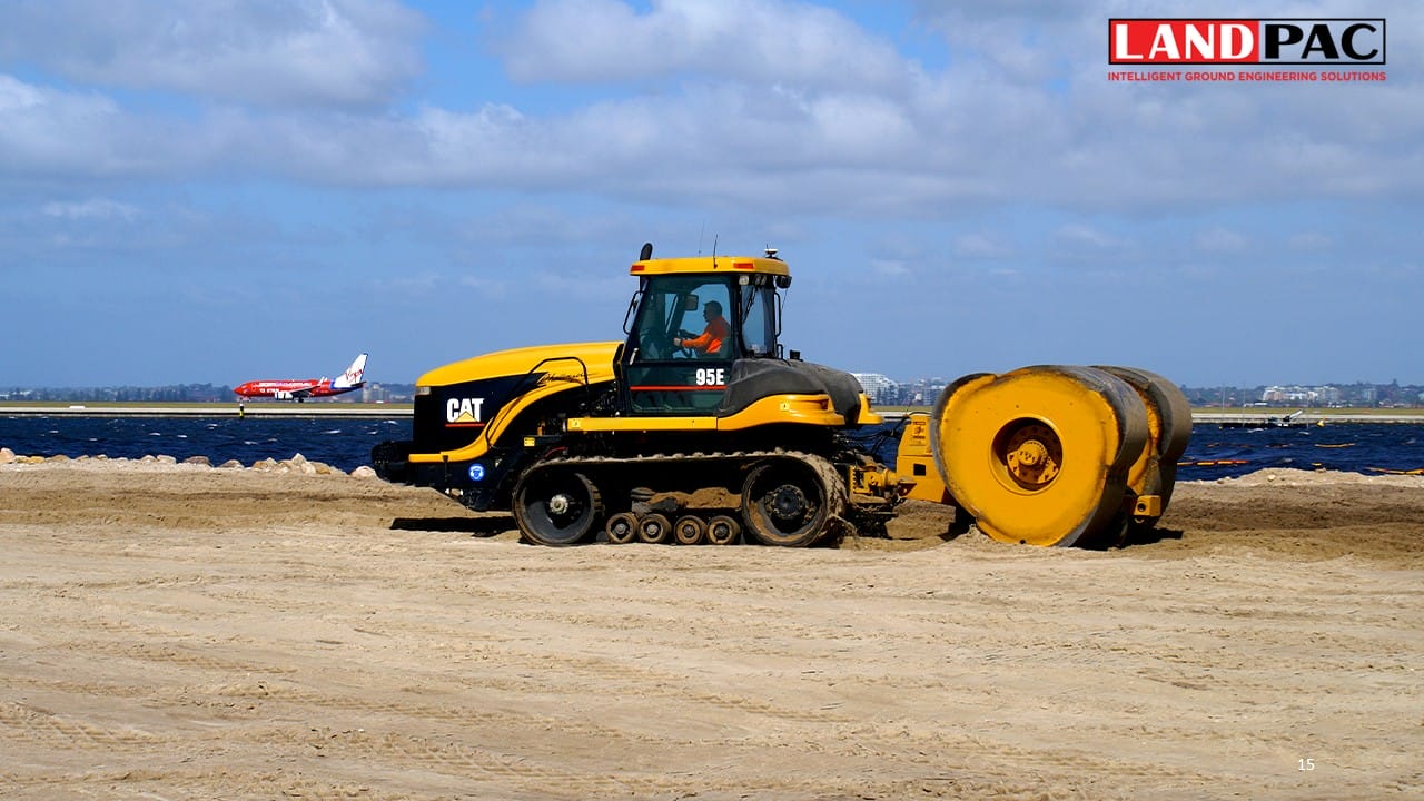 Yellow CAT track loader with roller on sandy site, plane in background, LANDPAC logo at top right.