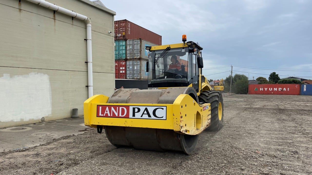 Yellow road roller labeled “LAND PAC” on gravel near stacked shipping containers.